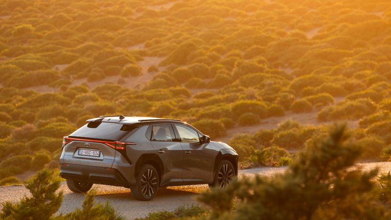 Toyota bZ4X gris vu de trois quarts arrière sur une route au coucher du soleil, dans un paysage naturel.