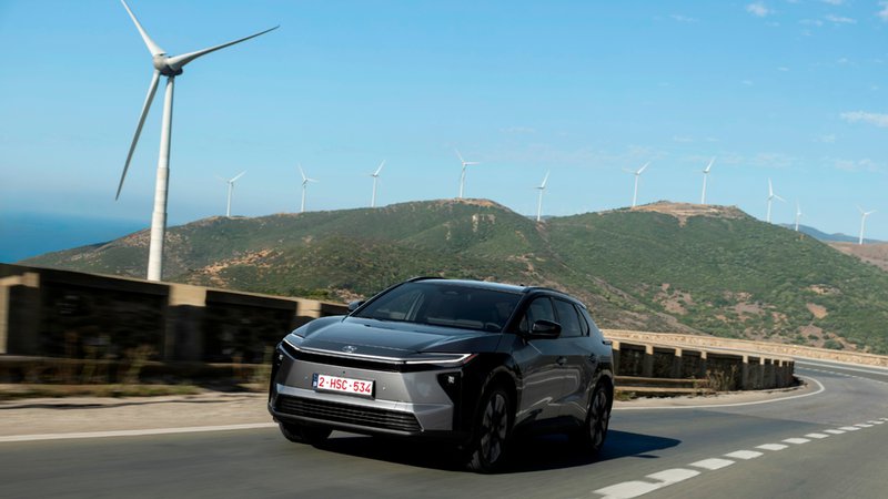 Toyota bZ4X gris vu de trois quarts avant en circulation sur une route de montagne, avec des éoliennes en arrière-plan.