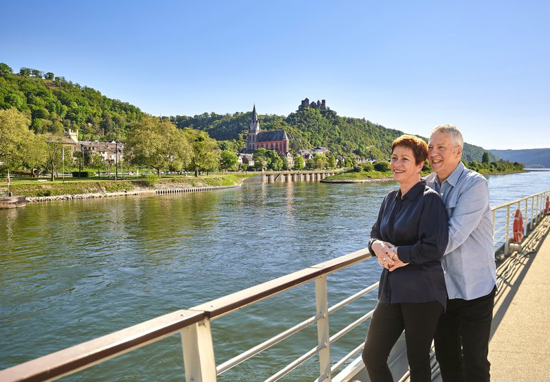 Koppel aan de reling van een schip op een rivier met groene oevers en een grote kerk, CroisiEurope
