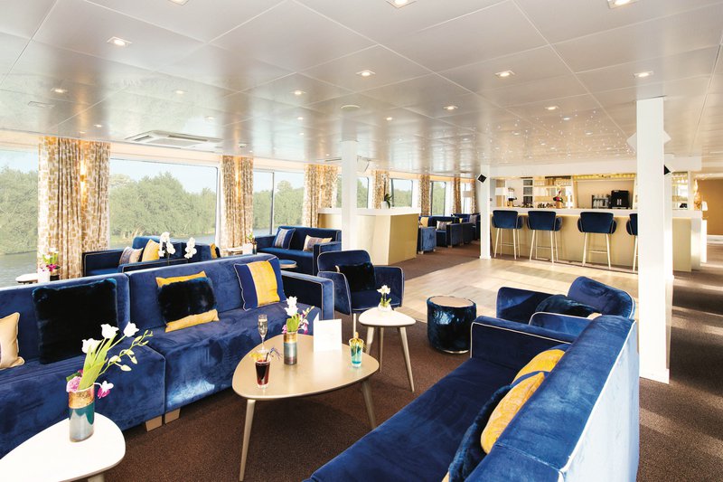 Interieur van een riviercruiseschip met blauwe zetels, grote ramen en een bar met vier krukken.