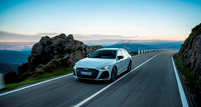Audi Audi break blanc roulant sur une route de montagne asphaltée avec montagnes et soleil couchant en arrière-plan