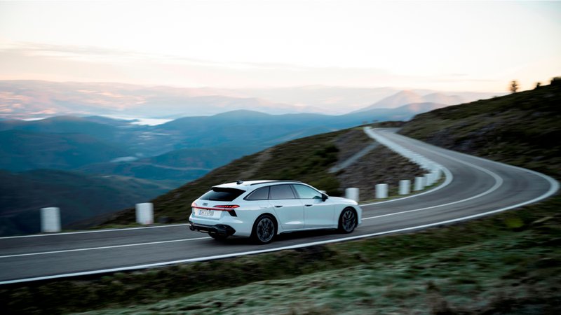 Audi break blanc roulant sur une route de montagne dans un virage avec montagnes et soleil couchant en arrière-plan