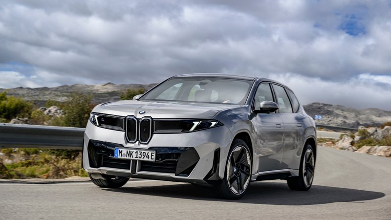 BMW iX3 gris vu de trois quarts avant, en mouvement sur une route de montagne sous un ciel chargé.