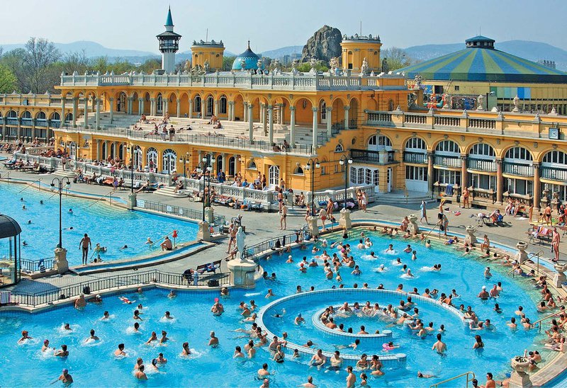 Des dizaines de personnes dans une grand piscine publique près d'un bâtiment majestueux.
