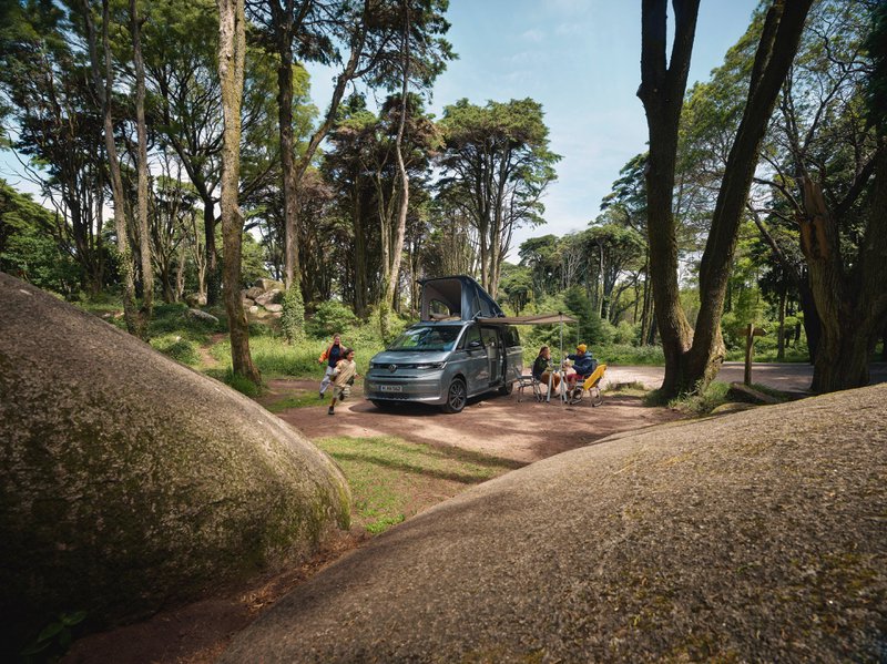 Un fourgon VW California entouré d'arbres et de personnes assis à une table de camping.