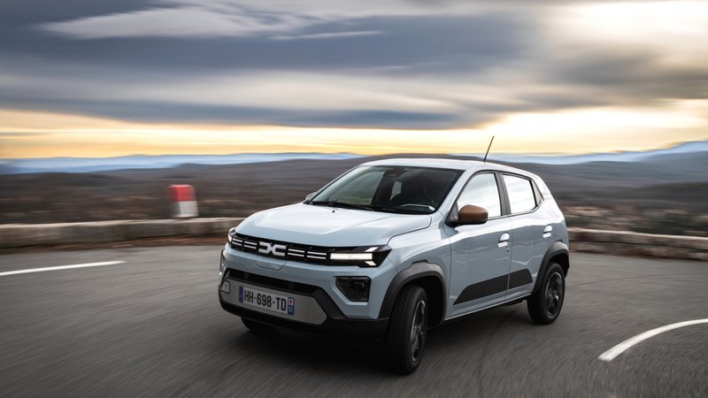 Dacia Spring électrique vue de trois quarts avant, en mouvement sur une route sinueuse au paysage dégagé.