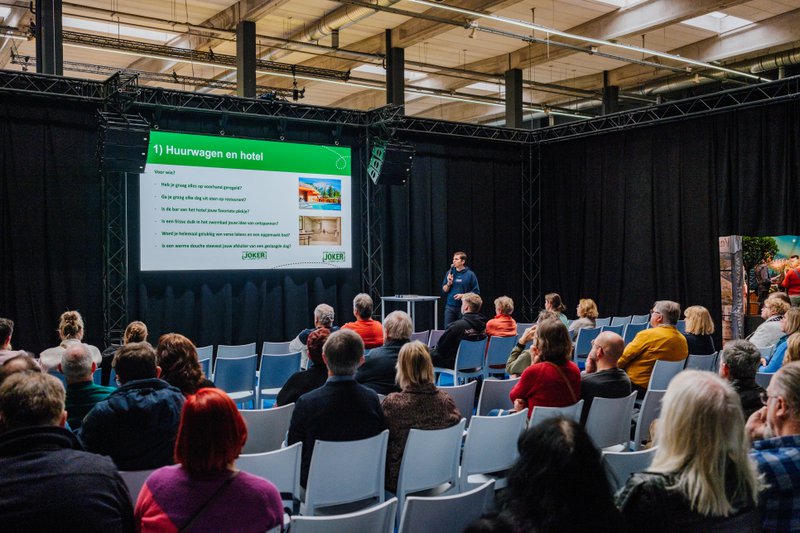 Mensen in een zaal volgen een presentatie op groot scherm. Vakantiebeurzen.