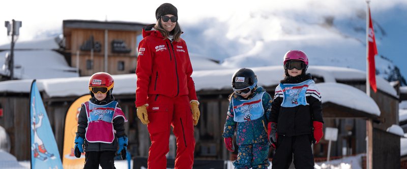 Monitrice de ski en tenue rouge avec 3 enfants devant un chalet de montagne.