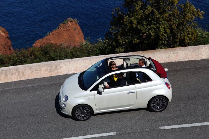 Vue aérienne sur une Fiat 500 blanche décapotée avec passagers, roulant sur une route ensoleillée surplombant la mer.
