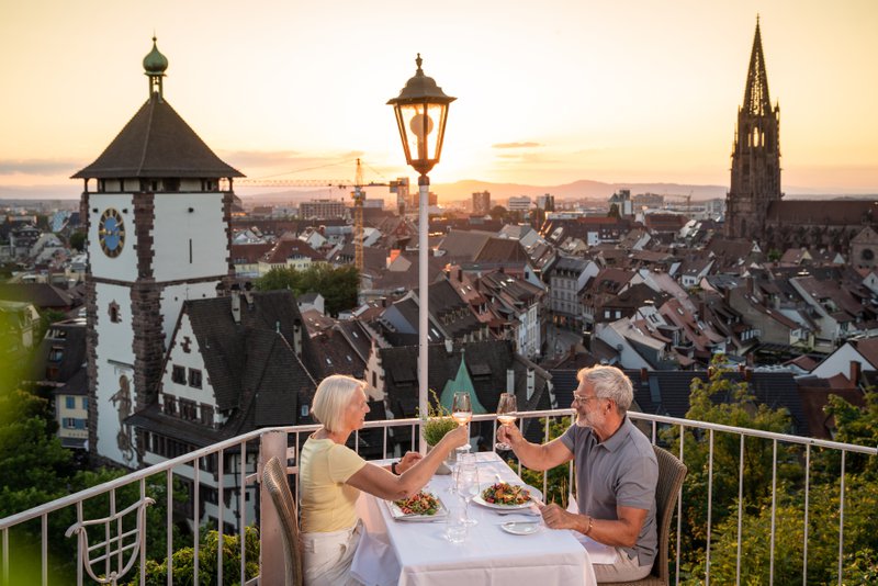 Un couple de personnes âgées déguste un verre de vin sur une terrasse au crépuscule avec vue sur la ville de Fribourg en Allemagne