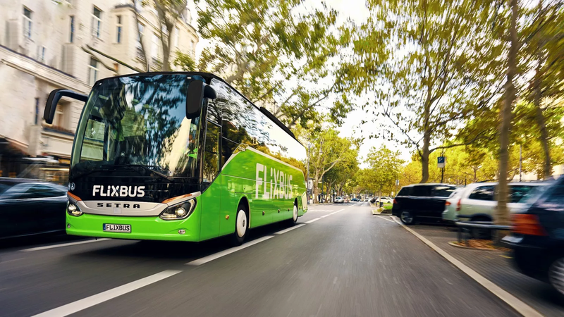Groene autocar op een tweevaksbaan geflankeerd door geparkeerde auto's.