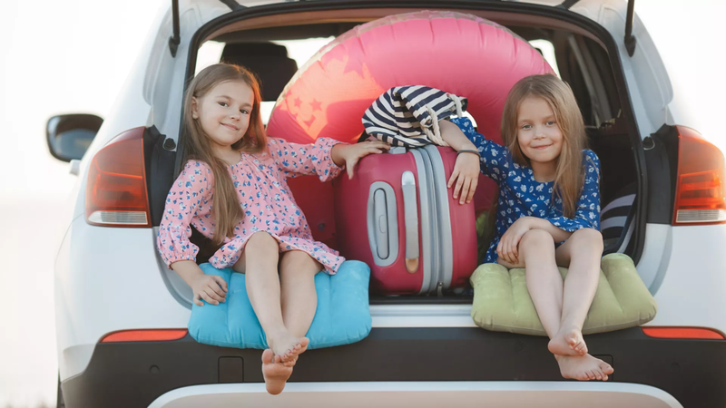 Twee kleine meisjes zitten tussen de bagage in de kofferruimte, en illustreren zo hoe belangrijk het is om je auto slim in te laden voor je vertrekt.