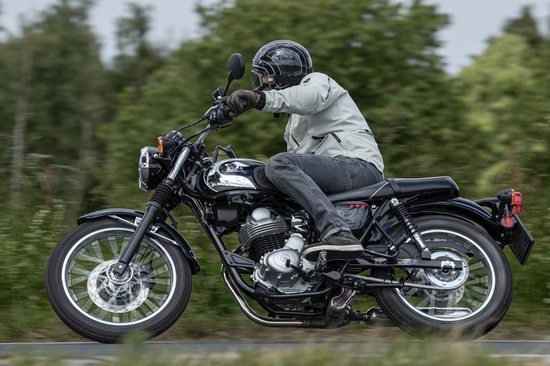 Zwarte Kawasaki Meguro S1 bestuurd door een motorrijder, met bomen op de achtergrond.