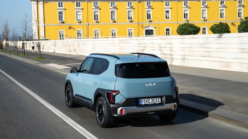 Kia EV2 bleu clair vue de trois quarts arrière, en mouvement sur une avenue urbaine.