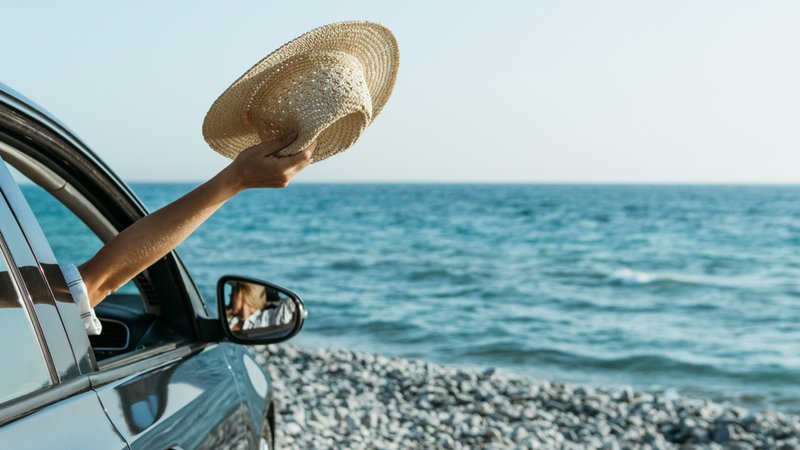Pause sur la route des vacances : une main tient un chapeau à la fenêtre d’une voiture arrêtée en bord de mer, face à la plage