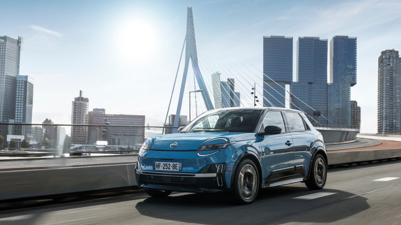 Nissan Micra bleue en mouvement sur une voie urbaine, avec skyline moderne et pont en arrière-plan.