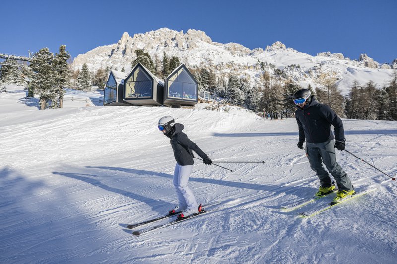 Un enfant skie avec un adulte dans un paysage hivernal au soleil avec une maison en verre en arrière-plan.