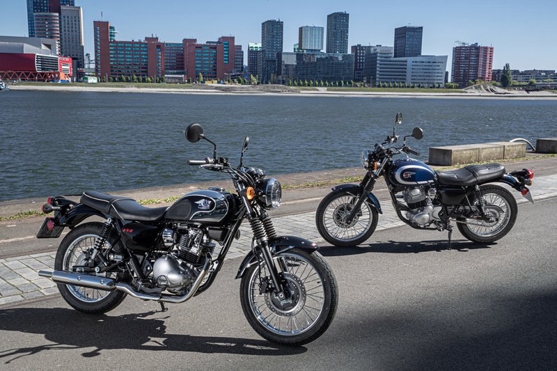 Twee Kawasaki Meguro's op een weg, met een rivier en moderne gebouwen op de achtergrond.
