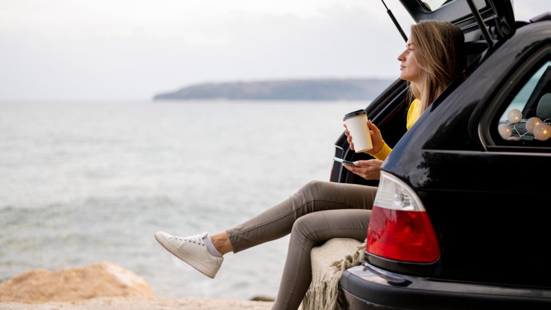 Une jeune femme assise à l'arrière du coffre de sa voiture, fait une pause près de la mer.