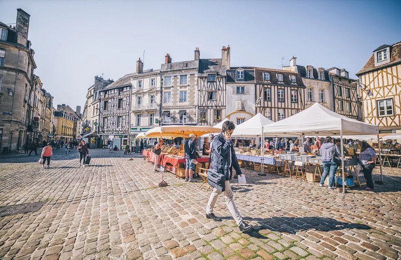 Place pavée bordée de maisons anciennes à colombages, avec des étalages de marché à droite et  à l'avant-plan un homme en train de téléphoner en marchant.