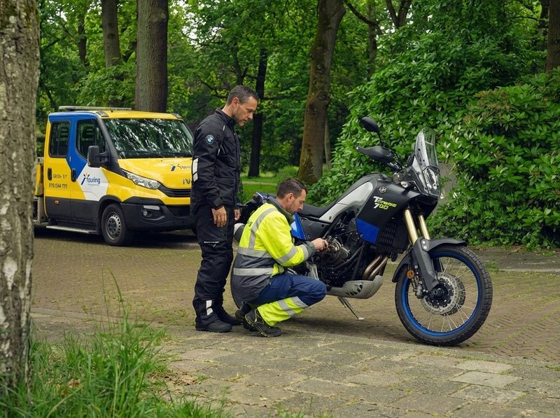 Un dépanneur contrôle une moto en panne sous le regard du motard sur une route boisée, camionnette Touring à l'arrière-plan.