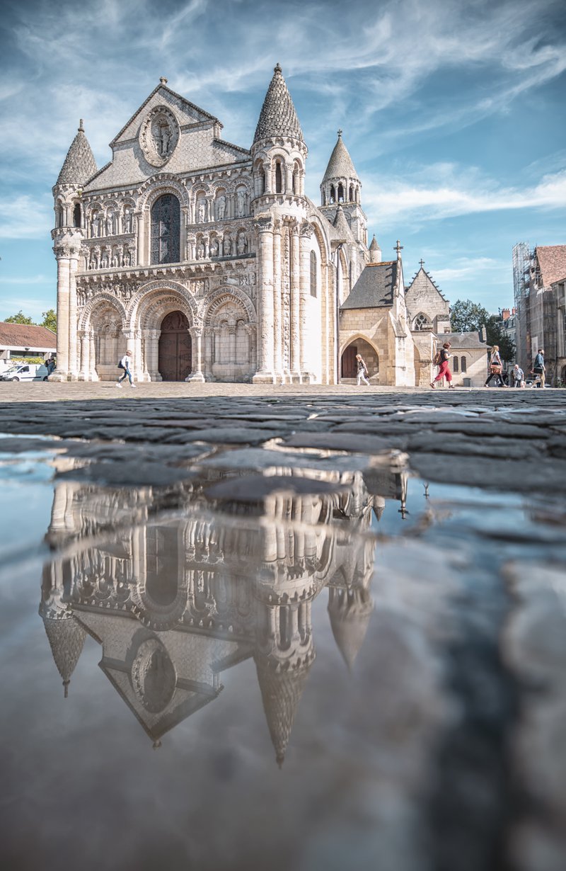 Façade d'une église romane ensoleillée avec son reflet dans une flaque d'eau, ciel bleu à peine voilé en arrière-plan.