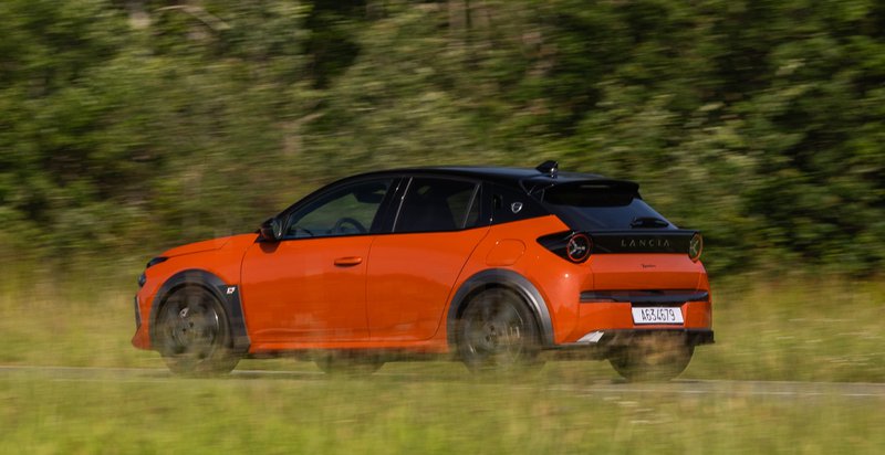 Vue arrière trois-quarts de la Lancia Ypsilon HF orange en mouvement sur route bordée de verdure.